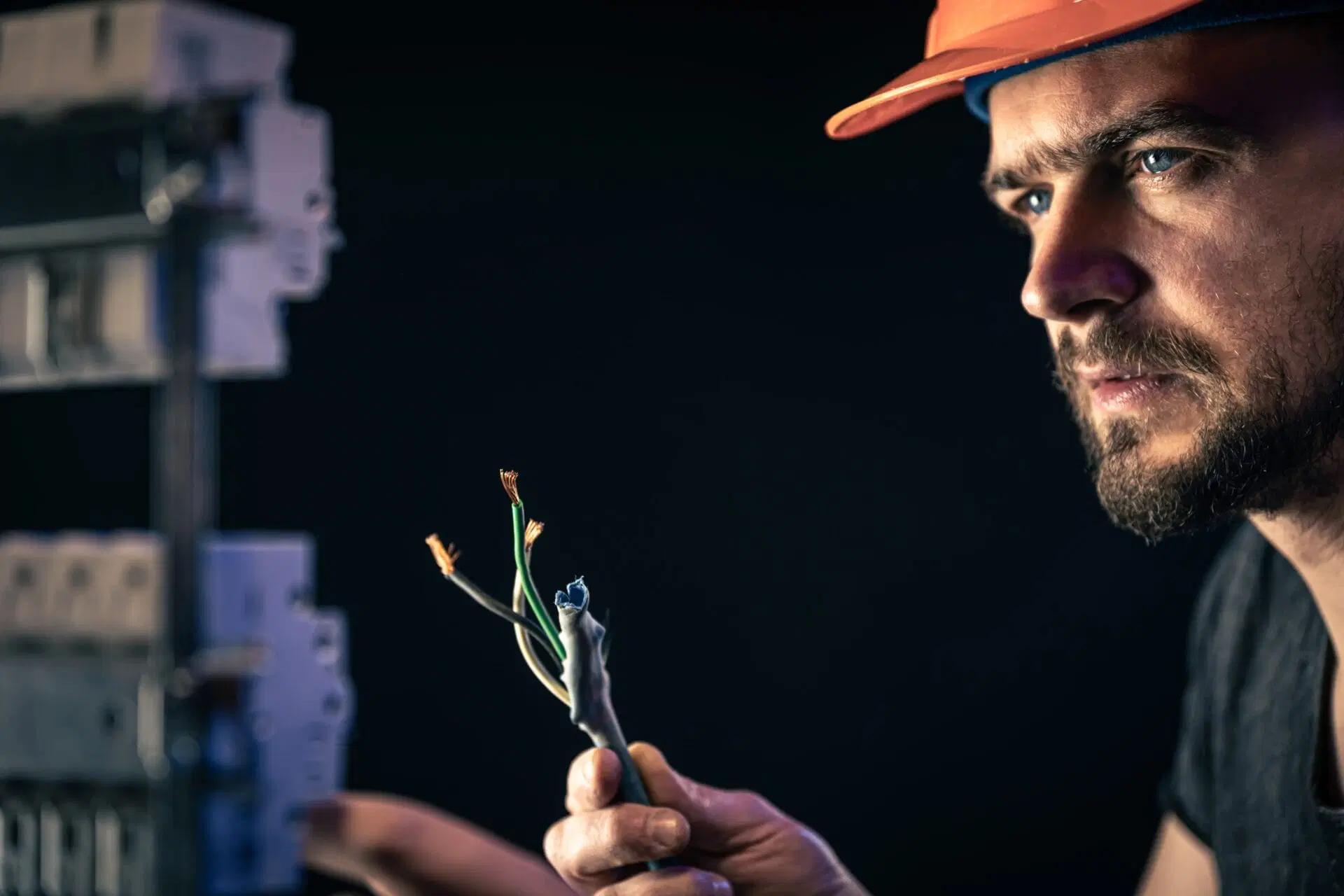 male-electrician-works-switchboard-with-electrical-connecting-cable-1-min Electrician wearing a hard hat inspecting exposed electrical wires with focused attention
